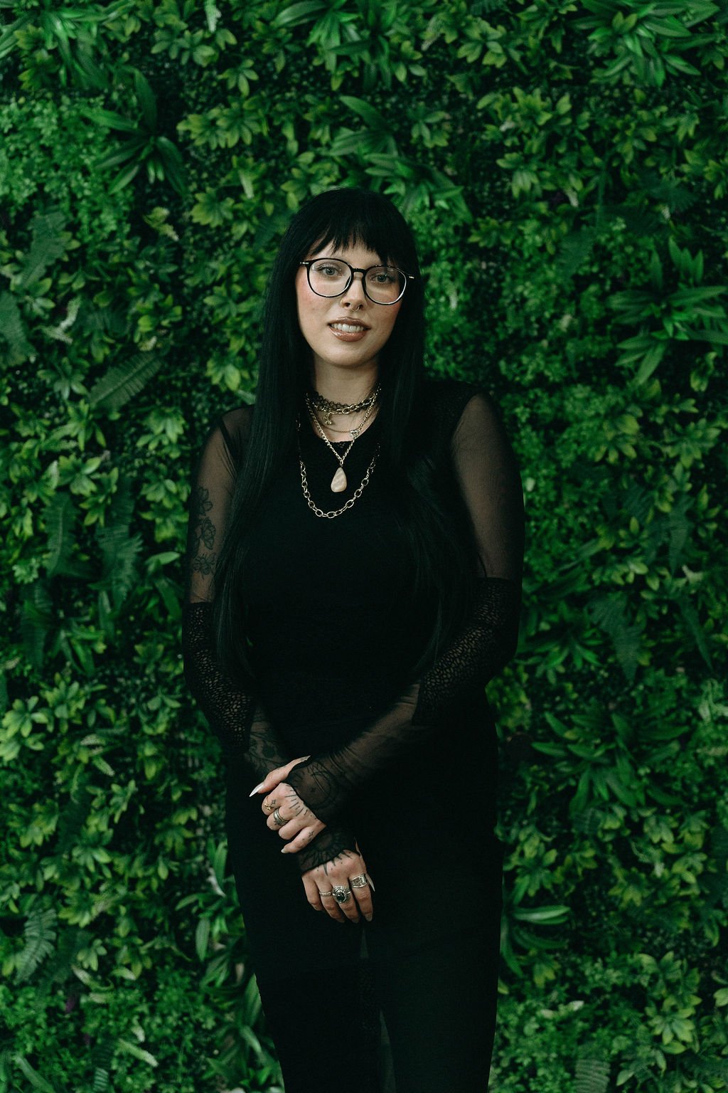Kelly wearing black and glasses, standing in front of a green foliage wall at Splat.